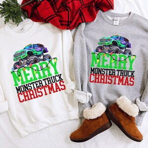 Op de afbeelding: Twee sweatshirts, één wit en één grijs, tonen een monster truck-ontwerp met de tekst "MERRY MONSTER TRUCK CHRISTMAS". De monstertruck is groen en paars. De tekst is groen en rood. Bruine laarzen met imitatiebont zijn op de voorgrond.