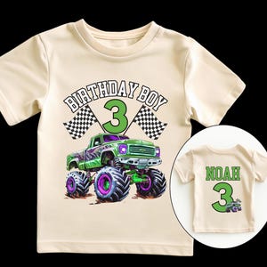 May include: Cream-colored t-shirt featuring a green monster truck graphic with purple and black wheels. The text "BIRTHDAY BOY 3" is above the truck, with checkered flags on either side. A smaller shirt shows the name "NOAH" and the number "3".