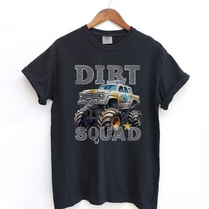 Puede incluir: Camiseta negra con un gr&aacute;fico de monster truck y la inscripci&oacute;n "DIRT SQUAD" en blanco a rayas. El cami&oacute;n es blanco y tiene neum&aacute;ticos negros grandes. La camiseta est&aacute; colgada en una percha de madera.