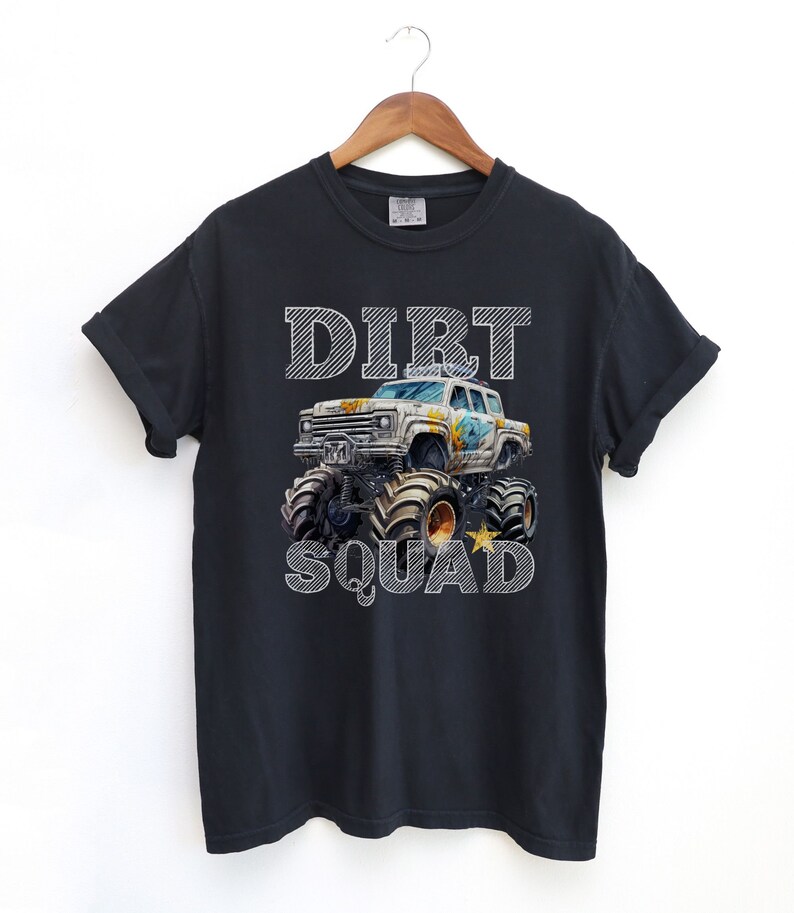 Puede incluir: Camiseta negra con un gr&aacute;fico de monster truck y la inscripci&oacute;n "DIRT SQUAD" en blanco a rayas. El cami&oacute;n es blanco y tiene neum&aacute;ticos negros grandes. La camiseta est&aacute; colgada en una percha de madera.