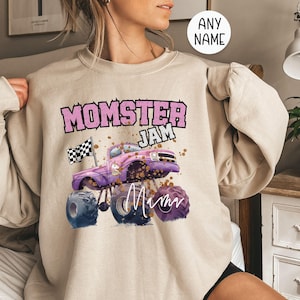 Pu&ograve; includere: Una felpa beige con una grafica di un monster truck rosa e viola e il testo "Momster Jam" e "Mom".