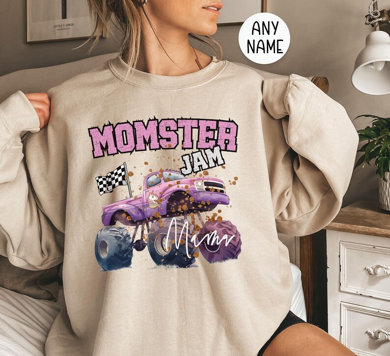 Pu&ograve; includere: Una felpa beige con una grafica di un monster truck rosa e viola e il testo "Momster Jam" e "Mom".
