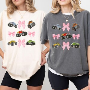 K&ouml;nnte beinhalten: Zwei &uuml;bergro&szlig;e T-Shirts, eines in Creme und eines in Anthrazit, jeweils mit einem sich wiederholenden Muster aus bunten Monstertrucks und rosa Schleifen. Die Trucks sind in verschiedenen Farben gehalten, darunter Blau, Orange, Rot, Gelb und Gr&uuml;n.
