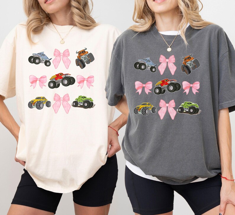 K&ouml;nnte beinhalten: Zwei &uuml;bergro&szlig;e T-Shirts, eines in Creme und eines in Anthrazit, jeweils mit einem sich wiederholenden Muster aus bunten Monstertrucks und rosa Schleifen. Die Trucks sind in verschiedenen Farben gehalten, darunter Blau, Orange, Rot, Gelb und Gr&uuml;n.
