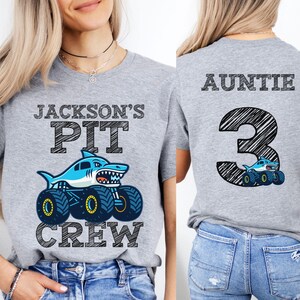 Op de afbeelding: Een lichtgrijs t-shirt met een haaienmonster truck ontwerp. De voorkant van het shirt leest "JACKSON'S PIT CREW" met een blauwe haai op een monstertruck. De achterkant van het shirt zegt "AUNTIE 3".