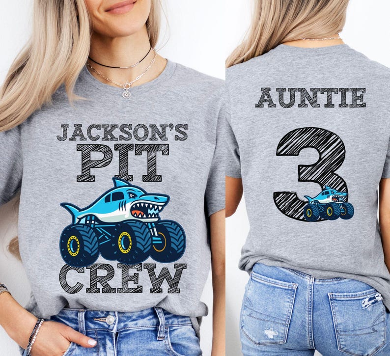 Op de afbeelding: Een lichtgrijs t-shirt met een haaienmonster truck ontwerp. De voorkant van het shirt leest "JACKSON'S PIT CREW" met een blauwe haai op een monstertruck. De achterkant van het shirt zegt "AUNTIE 3".