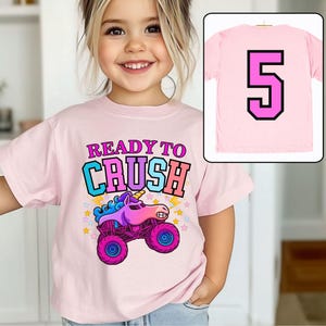 Pode incluir: T-shirt rosa claro com a frase "READY TO CRUSH" e um desenho de monster truck com cabe&ccedil;a de unic&oacute;rnio. A parte de tr&aacute;s da camiseta mostra o n&uacute;mero 5.