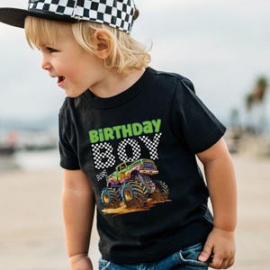 May include: Black t-shirt with a colorful monster truck graphic and the words "BIRTHDAY BOY" in green and checkered font. The shirt is worn by a child wearing a checkered hat.