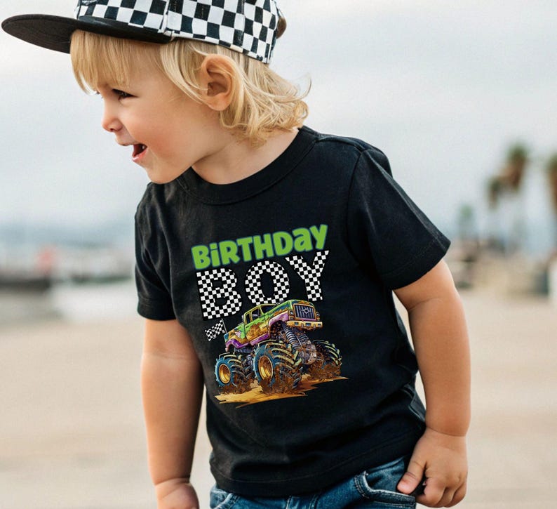 May include: Black t-shirt with a colorful monster truck graphic and the words "BIRTHDAY BOY" in green and checkered font. The shirt is worn by a child wearing a checkered hat.