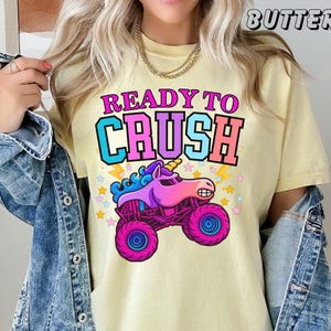 May include: Yellow t-shirt with the text "READY TO CRUSH" in pink, blue, and orange. The graphic features a cartoon unicorn monster truck with pink and purple wheels and a yellow horn. The word "BUTTER" is printed at the top.