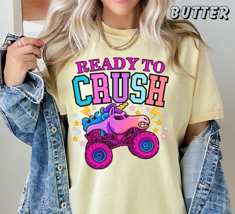 May include: Yellow t-shirt with the text "READY TO CRUSH" in pink, blue, and orange. The graphic features a cartoon unicorn monster truck with pink and purple wheels and a yellow horn. The word "BUTTER" is printed at the top.
