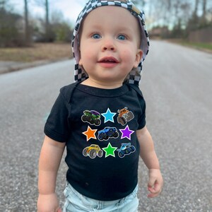 May include: A baby wearing a black short-sleeved bodysuit with colorful monster truck graphics inside star shapes. The baby is also wearing a checkered hat.
