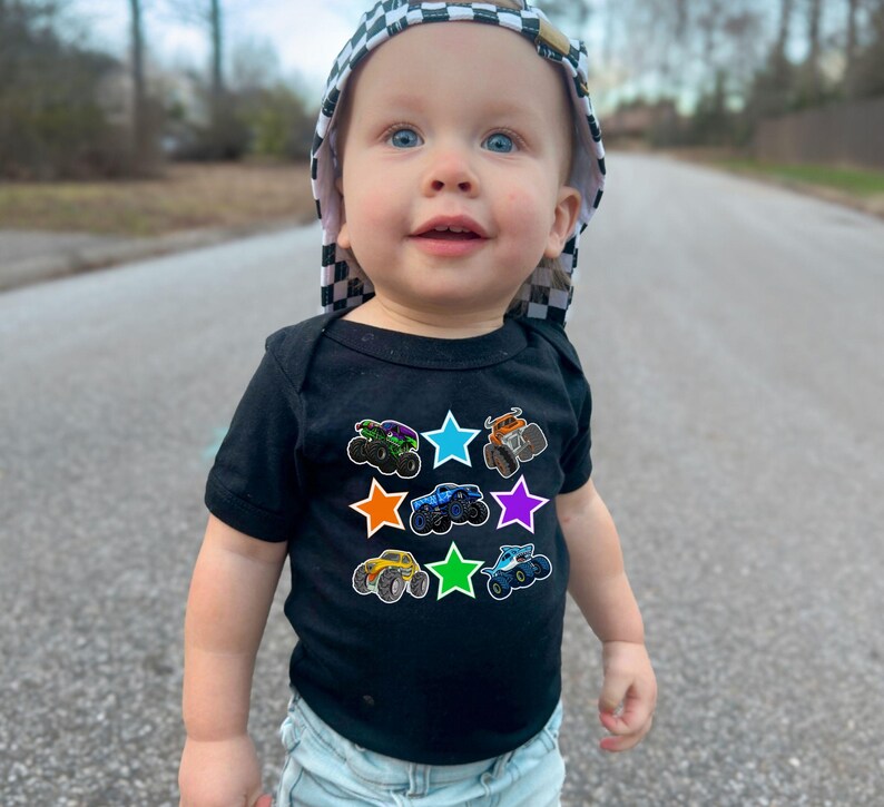 May include: A baby wearing a black short-sleeved bodysuit with colorful monster truck graphics inside star shapes. The baby is also wearing a checkered hat.