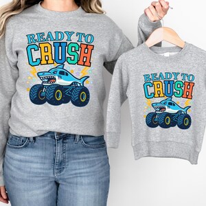 Pu&ograve; includere: Due felpe grigie, una per adulti e una per bambini, con un design di monster truck a forma di squalo. Il design include il testo "READY TO CRUSH" in blu, arancione e verde, con una grafica di squalo.