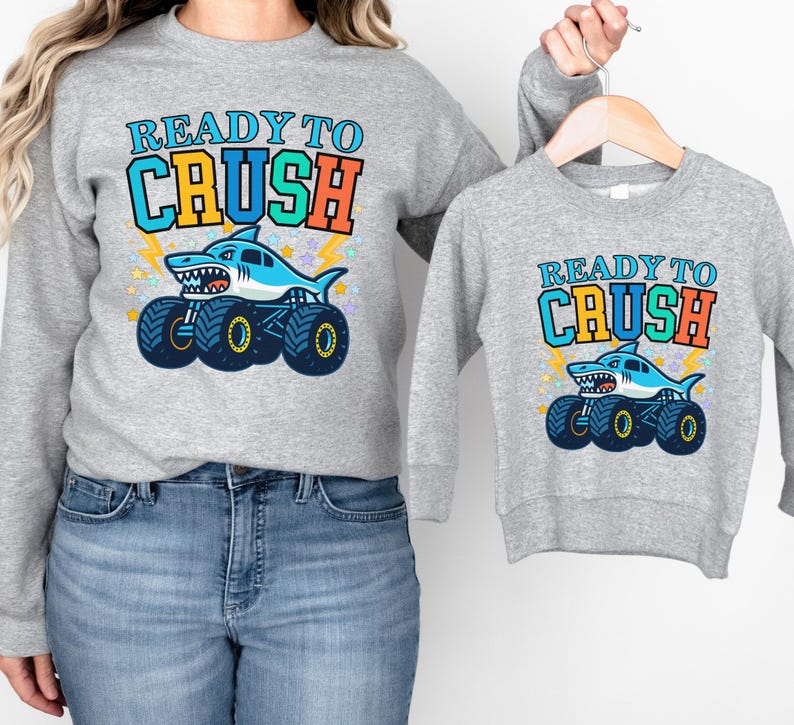 Pu&ograve; includere: Due felpe grigie, una per adulti e una per bambini, con un design di monster truck a forma di squalo. Il design include il testo "READY TO CRUSH" in blu, arancione e verde, con una grafica di squalo.