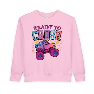 Puede incluir: Sudadera rosa claro con un diseño de camión monstruo unicornio y las palabras "READY TO CRUSH" en letras coloridas. El diseño incluye estrellas y rayos. La sudadera tiene mangas largas y cuello redondo.