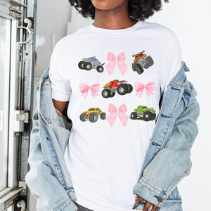 Op de afbeelding: Wit T-shirt met illustraties van kleurrijke monstertrucks en roze strikken. De monstertrucks zijn blauw, oranje, rood, geel en groen. De strikken zijn roze. Casual stijl.