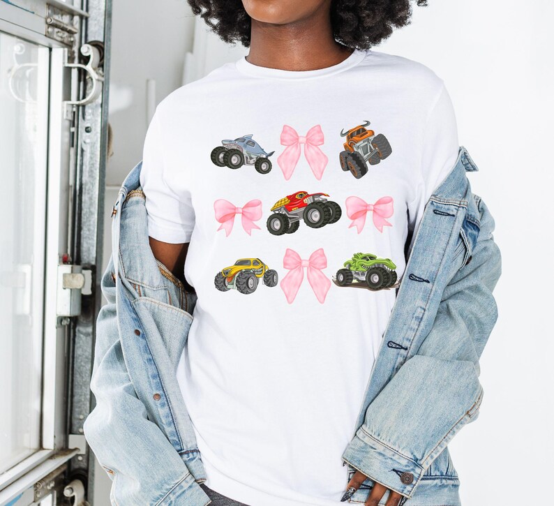 Op de afbeelding: Wit T-shirt met illustraties van kleurrijke monstertrucks en roze strikken. De monstertrucks zijn blauw, oranje, rood, geel en groen. De strikken zijn roze. Casual stijl.