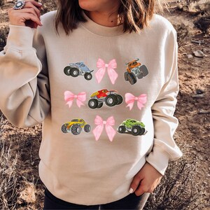 May include: Beige sweatshirt featuring colorful monster truck graphics and pink bows. The trucks are in various colors, including blue, orange, red, yellow, and green. The design is playful and suitable for casual wear.