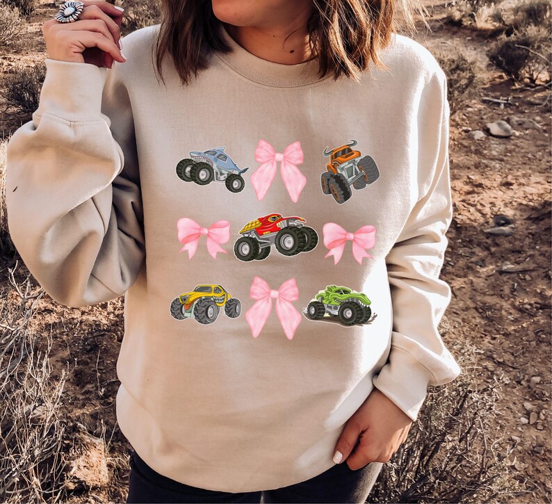 May include: Beige sweatshirt featuring colorful monster truck graphics and pink bows. The trucks are in various colors, including blue, orange, red, yellow, and green. The design is playful and suitable for casual wear.
