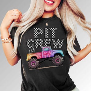 Woman wearing a Black t-shirt with the text "PIT CREW" in white and black checkered font. A colorful rainbow monster truck with a pink coquette bow flying a checkered flag  is below the text.