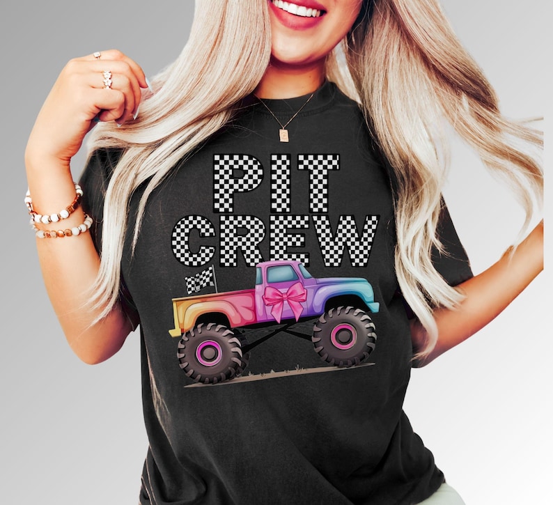 Woman wearing a Black t-shirt with the text "PIT CREW" in white and black checkered font. A colorful rainbow monster truck with a pink coquette bow flying a checkered flag  is below the text.