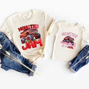 May include: Two cream-colored t-shirts with monster truck graphics. One shirt says "Momster Jam" with a red monster truck and strawberry-themed letters. The other says "Monster Trucks are my Jam" with a pink monster truck.