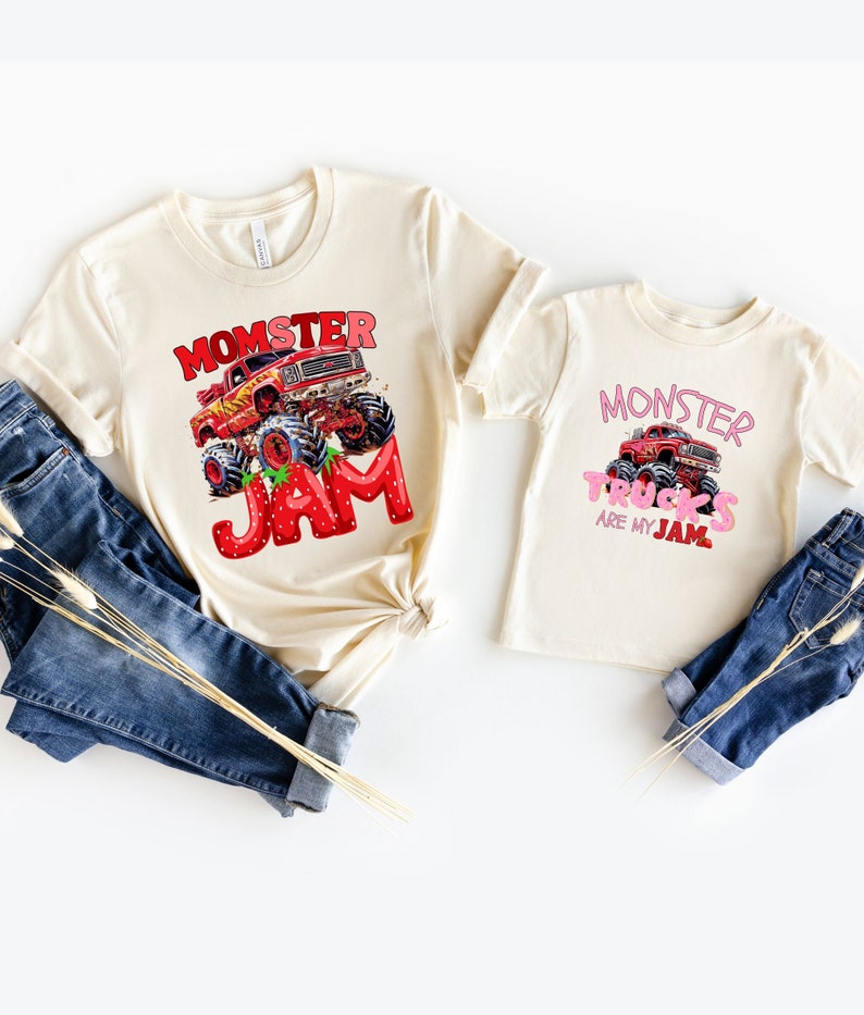 May include: Two cream-colored t-shirts with monster truck graphics. One shirt says "Momster Jam" with a red monster truck and strawberry-themed letters. The other says "Monster Trucks are my Jam" with a pink monster truck.