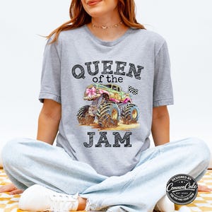 May include: A light grey t-shirt with a colourful monster truck graphic and the text "QUEEN of the JAM". The truck is green, pink, and blue, with large tyres and a chequered flag. Designed by Common Code.