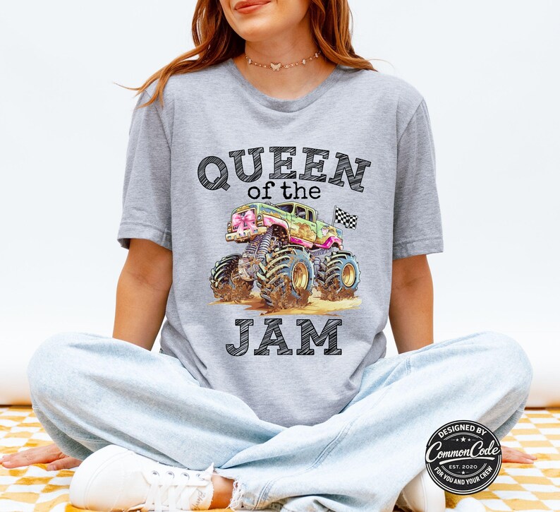 May include: A light grey t-shirt with a colourful monster truck graphic and the text "QUEEN of the JAM". The truck is green, pink, and blue, with large tyres and a chequered flag. Designed by Common Code.