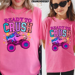 May include: Two pink t-shirts with the text "READY TO CRUSH" and a cartoon unicorn monster truck graphic. The graphic features a purple unicorn, blue mane, and pink wheels. The shirts are worn by two people.