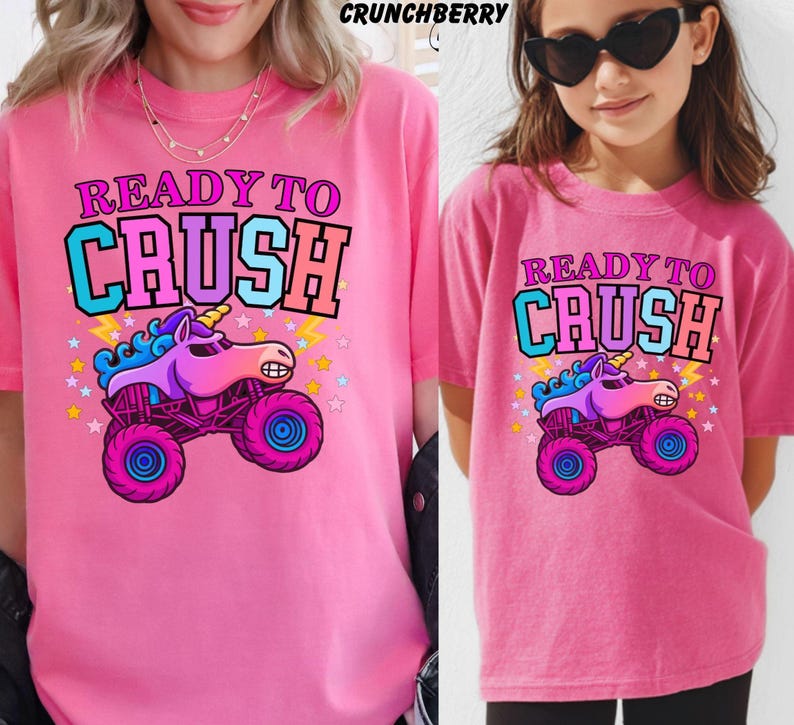 May include: Two pink t-shirts with the text "READY TO CRUSH" and a cartoon unicorn monster truck graphic. The graphic features a purple unicorn, blue mane, and pink wheels. The shirts are worn by two people.