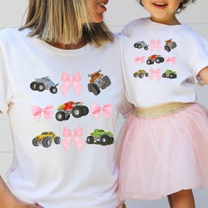 Puede incluir: Camisetas blancas con gráficos de monster trucks coloridos y detalles de lazos rosas. Una madre y su hijo llevan las camisetas. La niña también lleva una falda de tul rosa.