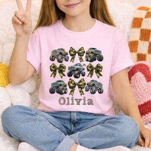 May include: A light pink t-shirt featuring a design of monster trucks and camouflage bows. The name "Olivia" is printed in a decorative font below the trucks. The shirt is paired with blue jeans.