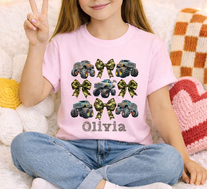 May include: A light pink t-shirt featuring a design of monster trucks and camouflage bows. The name "Olivia" is printed in a decorative font below the trucks. The shirt is paired with blue jeans.
