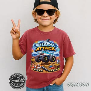 Boy is wearing a red Crimson Comfort Colors Monster Truck Jam tee with a Blue Megalodon Shark Truck that says "SHARK ATTACK" with a beach, pirate gold treasure, winners racing cup, ,checkered racing flags, This fun vibe is a great gift for any boy or man who loves sharks and monster big trucks. Race day truck show, great boy's birthday or Christmas gift, cool kid boy's shirt. Swimmer, Beach or pool party shirt
