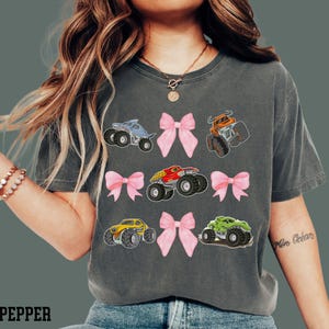 Woman model is wearing a Monster Truck Jam Theme t-shirt in Pepper by Comfort Colors. It is grid style placement of 4 croquette pink bows and 5 different monster trucks - Dragonoid, monster mutt, megalodon, El toro Loco, Dragon, that are Bull, Shark, Dog, Dinosaur themed trucks. Comes in Adult and Child Kid Sizes.