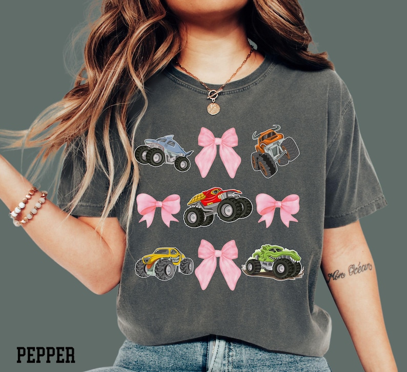 Woman model is wearing a Monster Truck Jam Theme t-shirt in Pepper by Comfort Colors. It is grid style placement of 4 croquette pink bows and 5 different monster trucks - Dragonoid, monster mutt, megalodon, El toro Loco, Dragon, that are Bull, Shark, Dog, Dinosaur themed trucks. Comes in Adult and Child Kid Sizes.