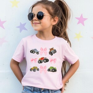 Op de afbeelding: Lichtroze T-shirt met kleurrijke monster truck afbeeldingen en roze strikaccenten. Het ontwerp toont monster trucks in blauw, rood, geel en groen. Het T-shirt heeft een ronde hals en korte mouwen.