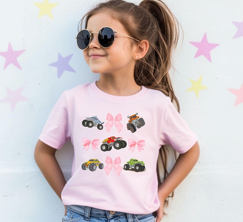Op de afbeelding: Lichtroze T-shirt met kleurrijke monster truck afbeeldingen en roze strikaccenten. Het ontwerp toont monster trucks in blauw, rood, geel en groen. Het T-shirt heeft een ronde hals en korte mouwen.