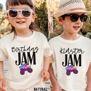 May include: Two children wearing cream-colored t-shirts with black text and a monster truck graphic. One shirt says "Birthday JAM" and the other says "Kidster JAM". The monster truck is pink, purple, and blue.