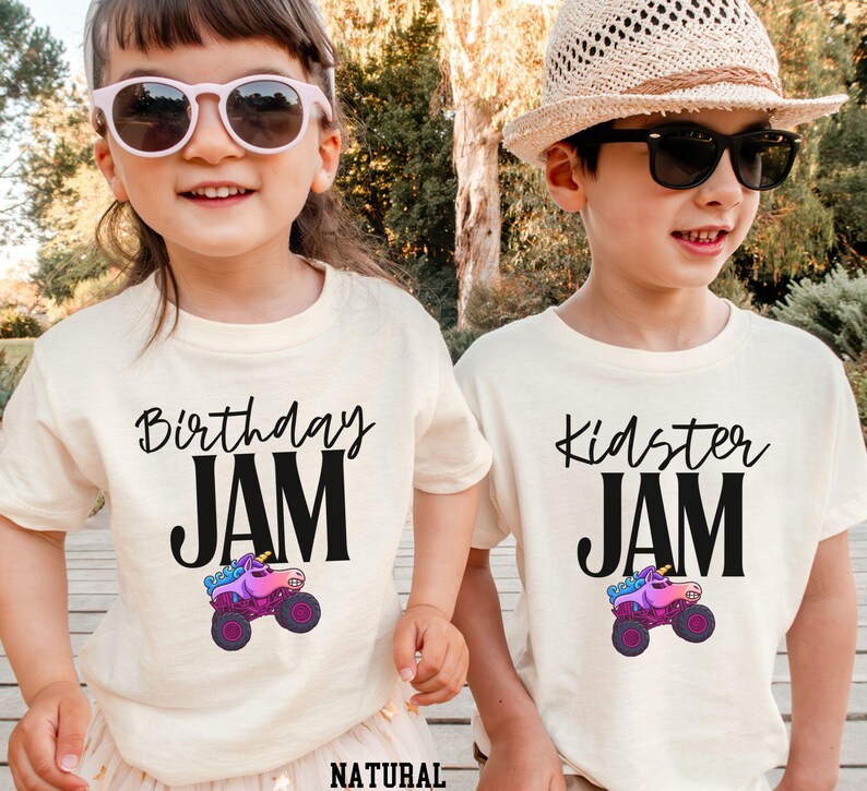 May include: Two children wearing cream-colored t-shirts with black text and a monster truck graphic. One shirt says "Birthday JAM" and the other says "Kidster JAM". The monster truck is pink, purple, and blue.