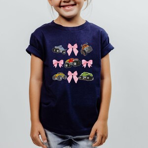 Op de afbeelding: Een marineblauw T-shirt met geborduurde monstertrucks en roze strikken. Het ontwerp bevat zes verschillende monstertruck illustraties in verschillende kleuren, elk gecombineerd met een roze strik. Het T-shirt wordt door een jong kind gedragen.