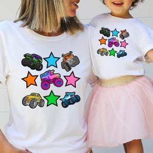Może przedstawiać: Białe koszulki z kolorową grafiką monster truck&oacute;w i gwiazdkami. Koszulki przedstawiają r&oacute;żne wzory monster truck&oacute;w w kolorach zielonym, pomarańczowym, r&oacute;żowym, ż&oacute;łtym i niebieskim. Koszulki noszą matka i dziecko.