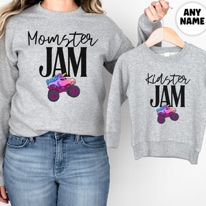 May include: Two gray sweatshirts, one adult and one child size, with matching designs. The adult sweatshirt says "Momster JAM" with a monster truck graphic. The child's sweatshirt says "Kidster JAM" with a similar graphic. The monster truck is pink, purple, and blue.