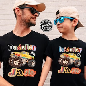 Puede incluir: Dos camisetas negras con gr&aacute;ficos coloridos. Una camiseta dice "Dadster Jam" con un dise&ntilde;o de cami&oacute;n monstruo amarillo. La otra dice "Kidster Jam" con un cami&oacute;n similar. Ambas camisetas tienen una bandera a cuadros.