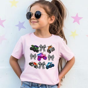 May include: A light pink t-shirt featuring colorful monster trucks and checkered bows. The monster trucks are in various colors, including green, orange, red, blue, and pink. The bows are black and white. The shirt is worn by a young girl.