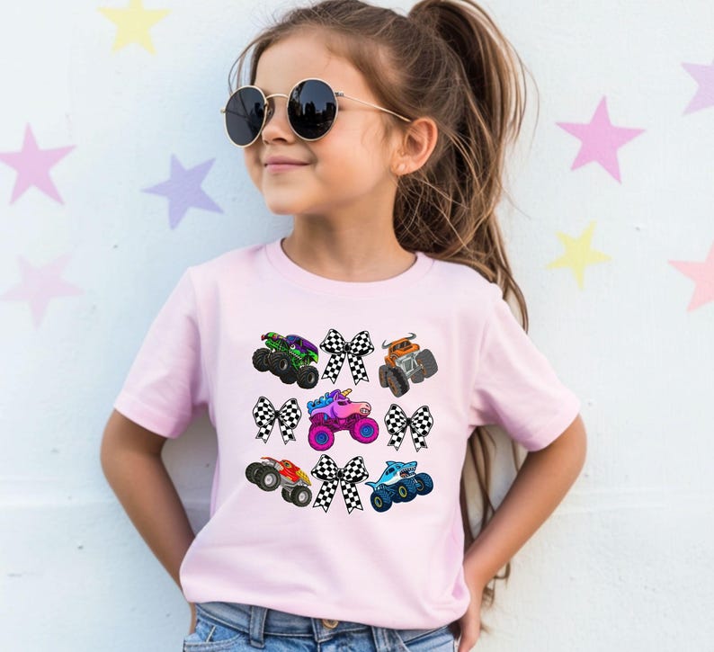 May include: A light pink t-shirt featuring colorful monster trucks and checkered bows. The monster trucks are in various colors, including green, orange, red, blue, and pink. The bows are black and white. The shirt is worn by a young girl.
