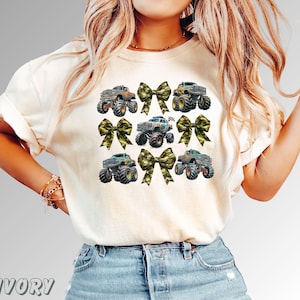 May include: Ivory-colored t-shirt featuring a design of monster trucks and camouflage bows. The trucks are in various colors, with large tires. The bows are in a camouflage pattern. The word "IVORY" is printed at the bottom.