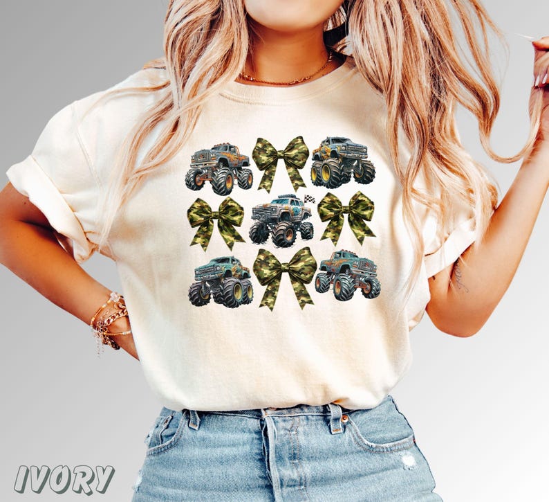 May include: Ivory-colored t-shirt featuring a design of monster trucks and camouflage bows. The trucks are in various colors, with large tires. The bows are in a camouflage pattern. The word "IVORY" is printed at the bottom.